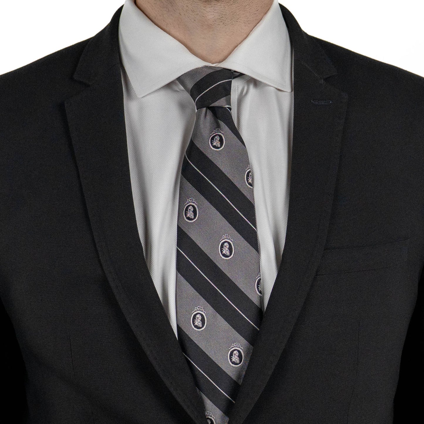 A close-up of a person wearing a black suit jacket and white dress shirt, with a gray tie featuring diagonal black stripes and small crest motifs.