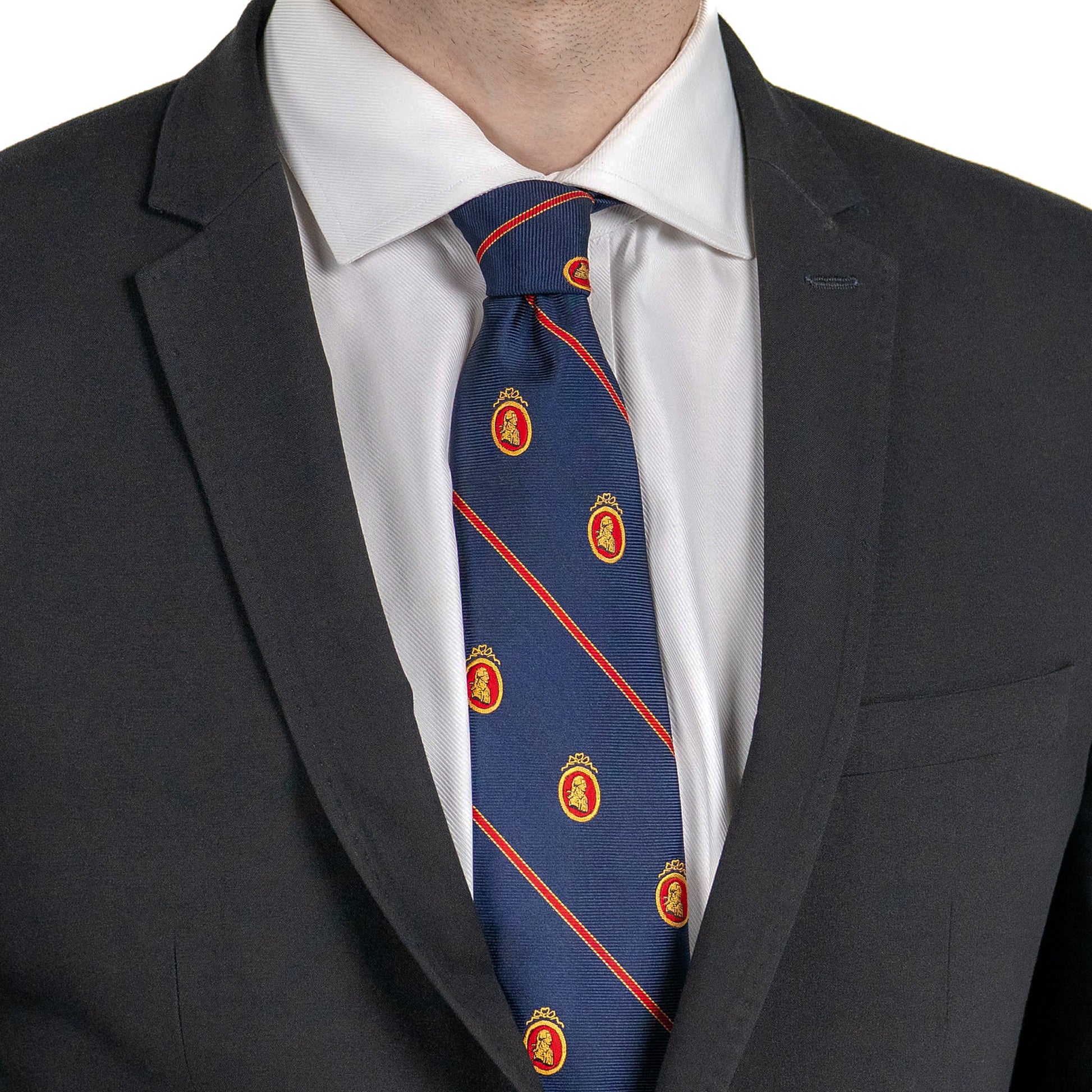 A close-up of a person wearing a dark suit jacket and white dress shirt, with a navy blue tie featuring diagonal red stripes and small gold-and-red crest patterns.