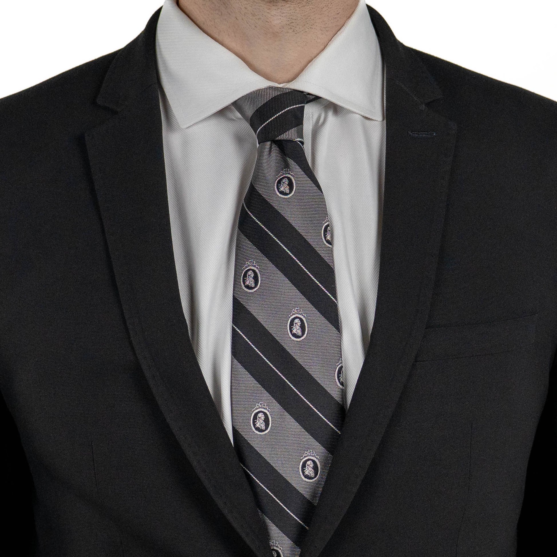 A close-up of a person wearing a black suit jacket and white dress shirt, with a gray tie featuring diagonal black stripes and small crest motifs.